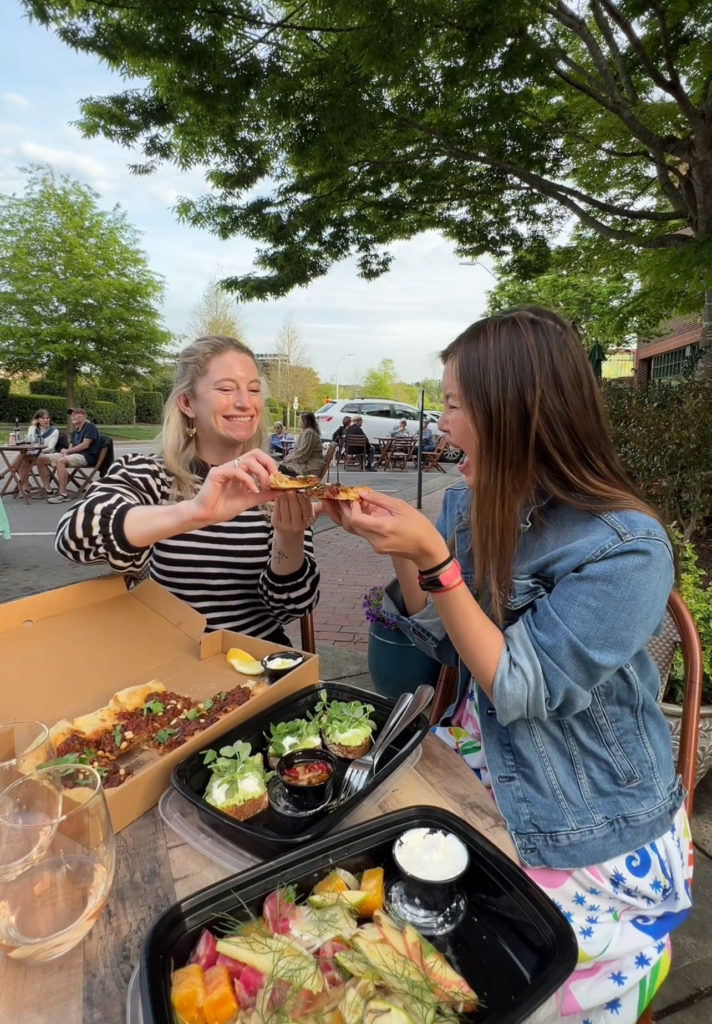 Photograph of blond woman and Asian woman cheersing flatbread while enjoying a meal alfresco at Wine Down Wednesday event in Chapel Hill