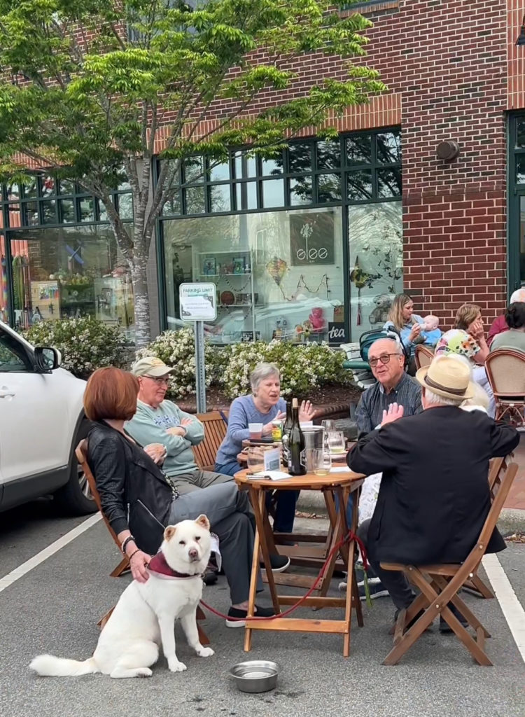 Group of 5-6 older people sitting outdoors with a table with wine bottles and a white dog at Wine Down Wednesday event in Chapel Hill
